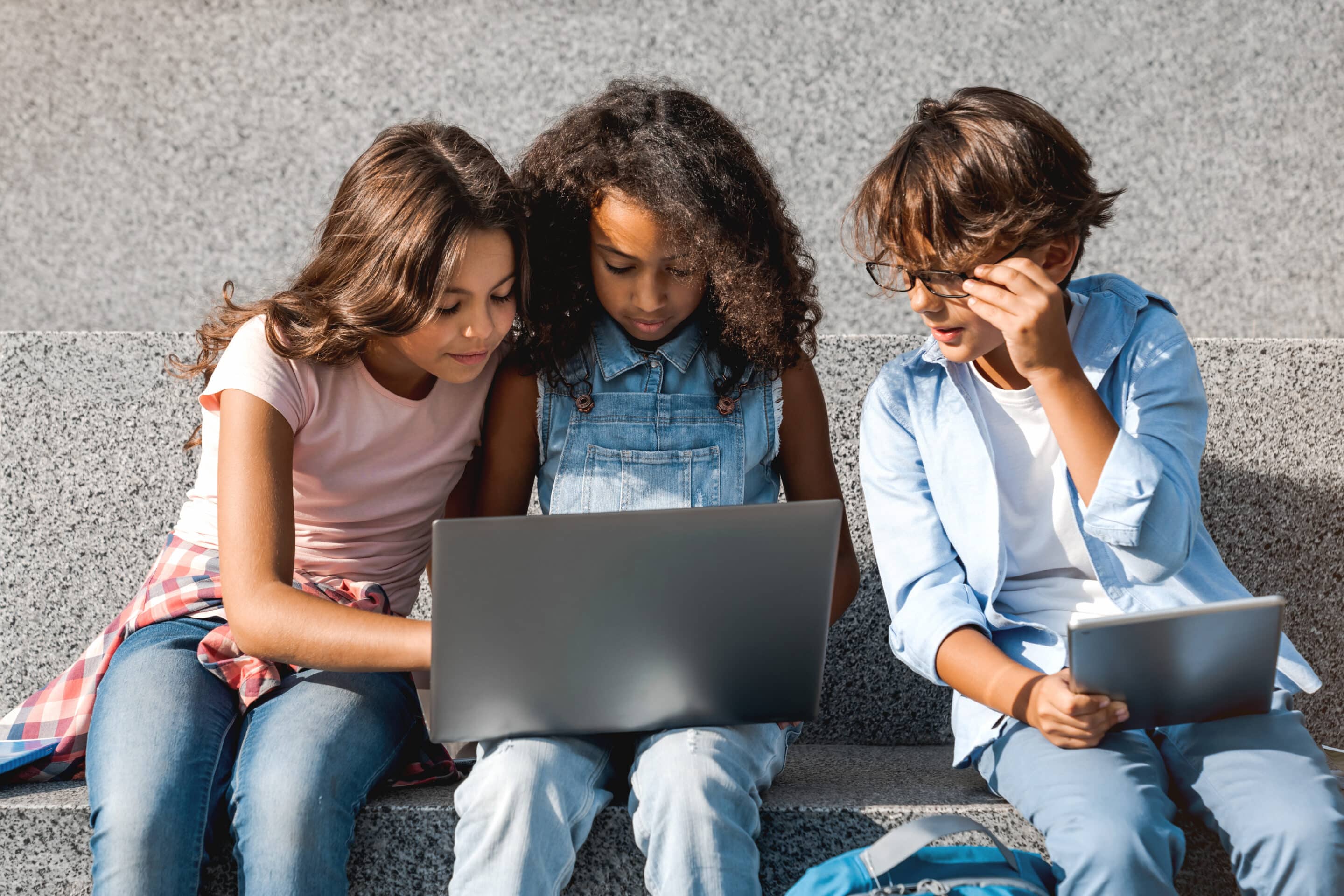 Smiling school kids sitting and looking in laptop screen with digital devices gadgets in hands sitting on the stairs outdoors. Elementary middle pupils doing home work together